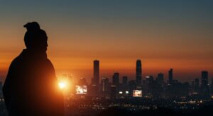 A dynamic, thought-provoking image blending ancient East Asian philosophy with modern urban life. In the foreground, a silhouetted figure of Mencius, perhaps with a subtle glow, stands contemplating a modern city skyline at dusk. The city should show elements of bustling activity and illuminated screens, representing 'profit' and 'competition.' Behind Mencius, gentle, warm light emanates, symbolizing 'benevolence' and 'righteousness.' The overall mood should be contemplative and hopeful, with a palette of deep terracotta, soft grays, and warm oranges, reflecting the chosen theme.